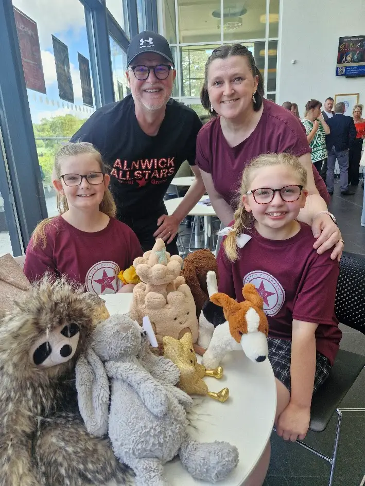 Family of four smiles behind a table piled with plush stuffed animals at an indoor event.