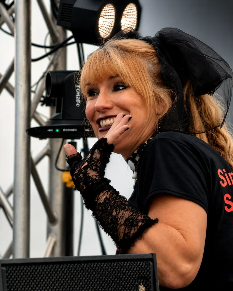 Smiling blonde female singer on stage, wearing a black lace glove and a large black bow, stage lights behind her.